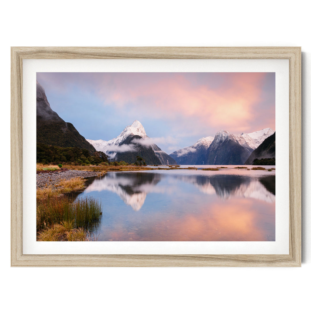 a serene landscape with a calm lake reflecting the surrounding mountains and sky. The mountains are covered in snow, and the sky is a beautiful gradient of pink and orange, creating a stunning backdrop for the scene.
