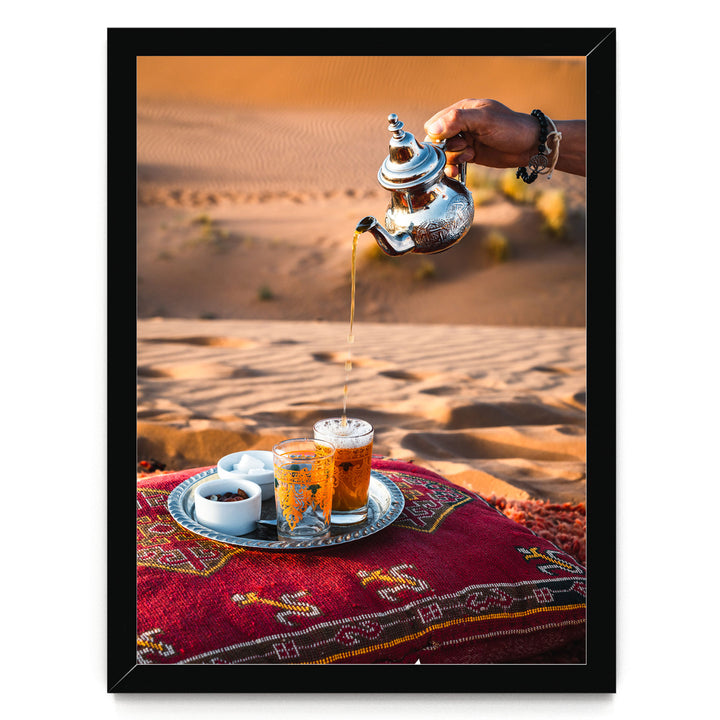 A hand pours tea from a silver teapot into two glasses on a red and gold patterned tablecloth, set against a desert landscape.
