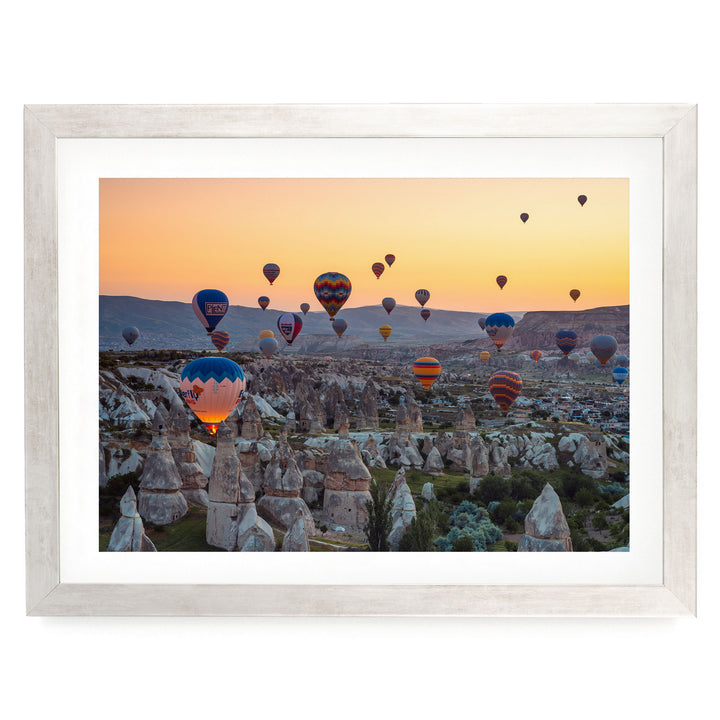 a breathtaking scene of hot air balloons floating over a mountainous landscape at sunset. The balloons are painted in a variety of vibrant colors, creating a stunning visual display against the backdrop of the rocky terrain and the warm hues of the setting sun.