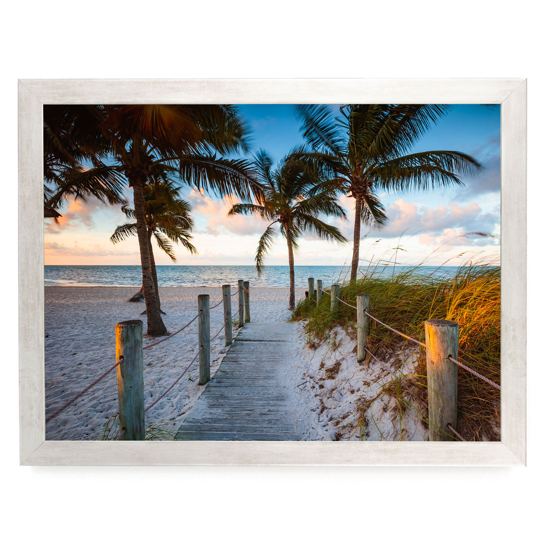 A wooden walkway leads to a sandy beach, with palm trees and a colorful sunset sky in the background.