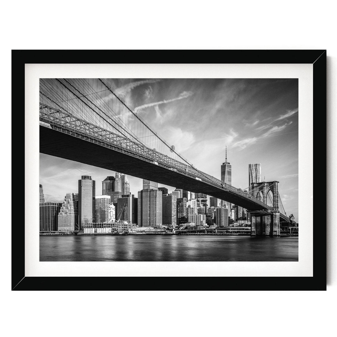 A black and white photograph of the Brooklyn Bridge spanning the East River, with the city skyline of New York City visible in the background.