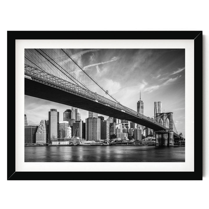 A black and white photograph of the Brooklyn Bridge spanning the East River, with the city skyline of New York City visible in the background.