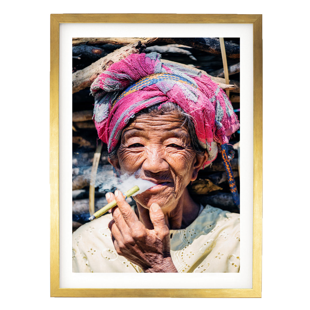 An elderly woman wearing a pink headscarf and a white blouse is shown smoking a cigarette, with a pile of wood in the background.
