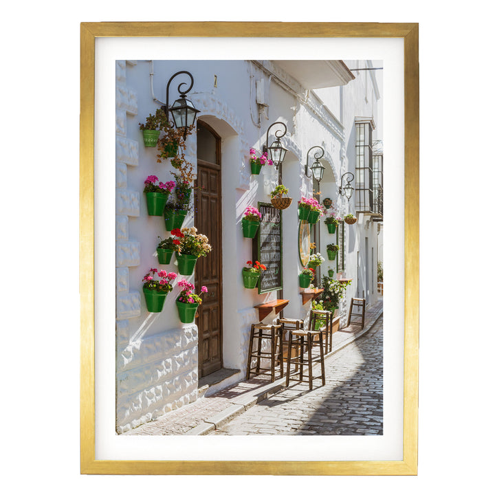 A charming street scene with a white building adorned with hanging flower pots and a wooden door. The building features a sign with a green and white color scheme. The street is lined with wooden chairs and tables, creating a cozy outdoor seating area.
