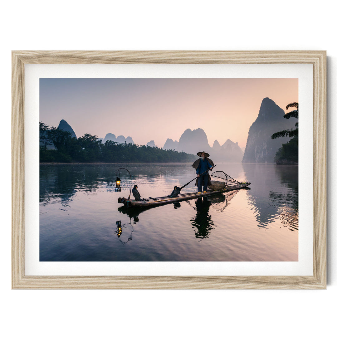 A person stands on a long wooden boat, paddling across a calm body of water surrounded by mountains and trees.