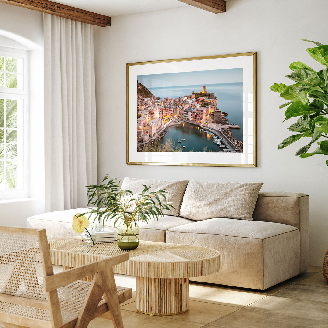 a living room with a large framed picture of a coastal town hanging on the wall. The room has a beige sofa, a wooden coffee table, and a potted plant. The overall decor has a modern and minimalist style.