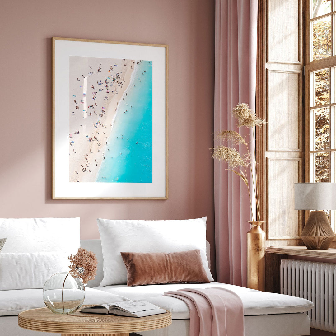 a cozy living room with a large window, a framed picture of a beach scene, a white sofa, a wooden coffee table, and a vase with dried flowers.
