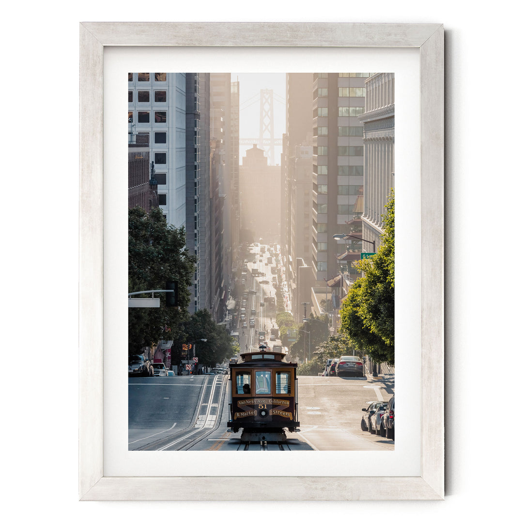 A framed photograph depicting a trolley car traveling down a city street, with tall buildings and trees lining the street.