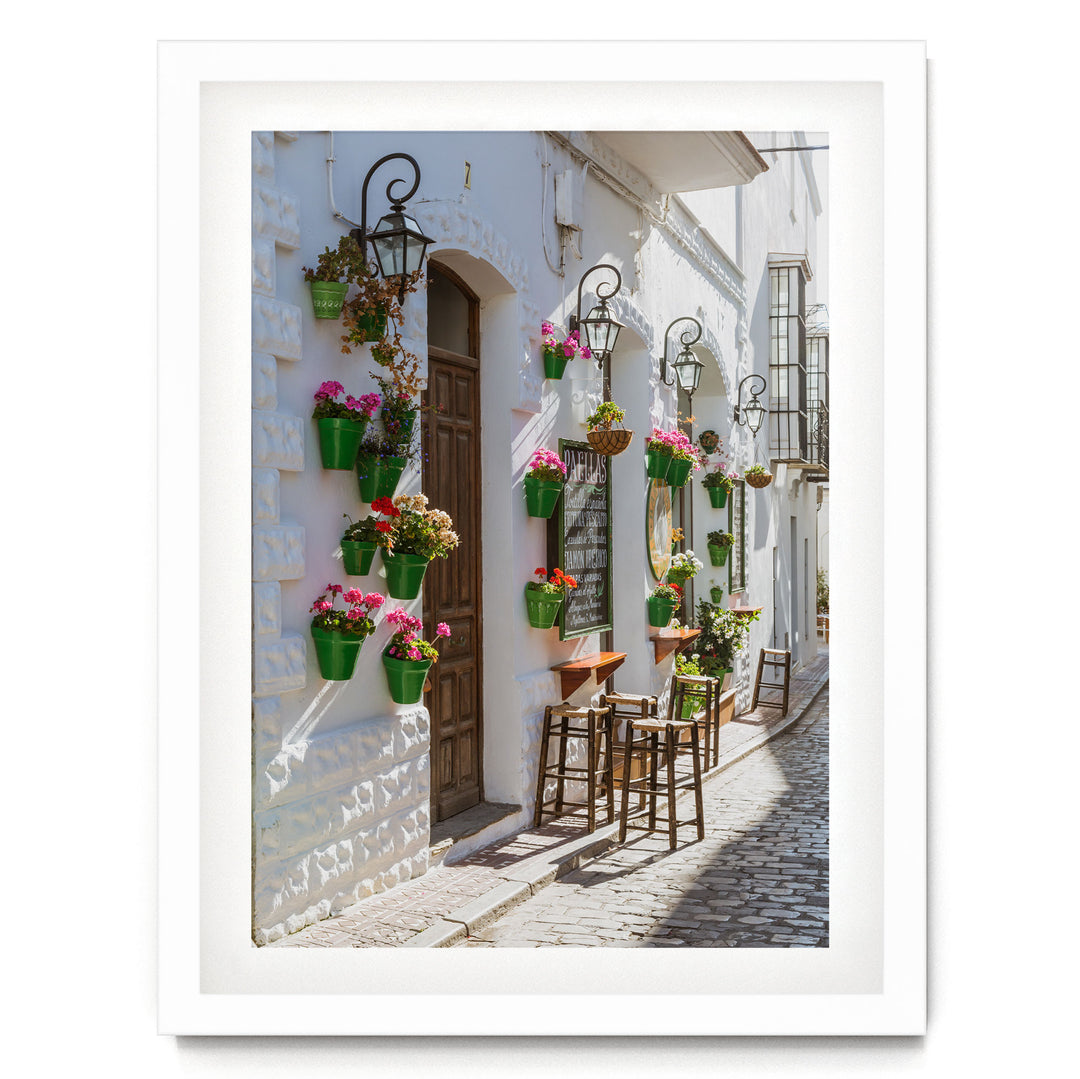 A charming street scene with a white building adorned with hanging flower pots and a wooden door. The building features a small balcony with a table and chairs, creating a cozy and inviting atmosphere.