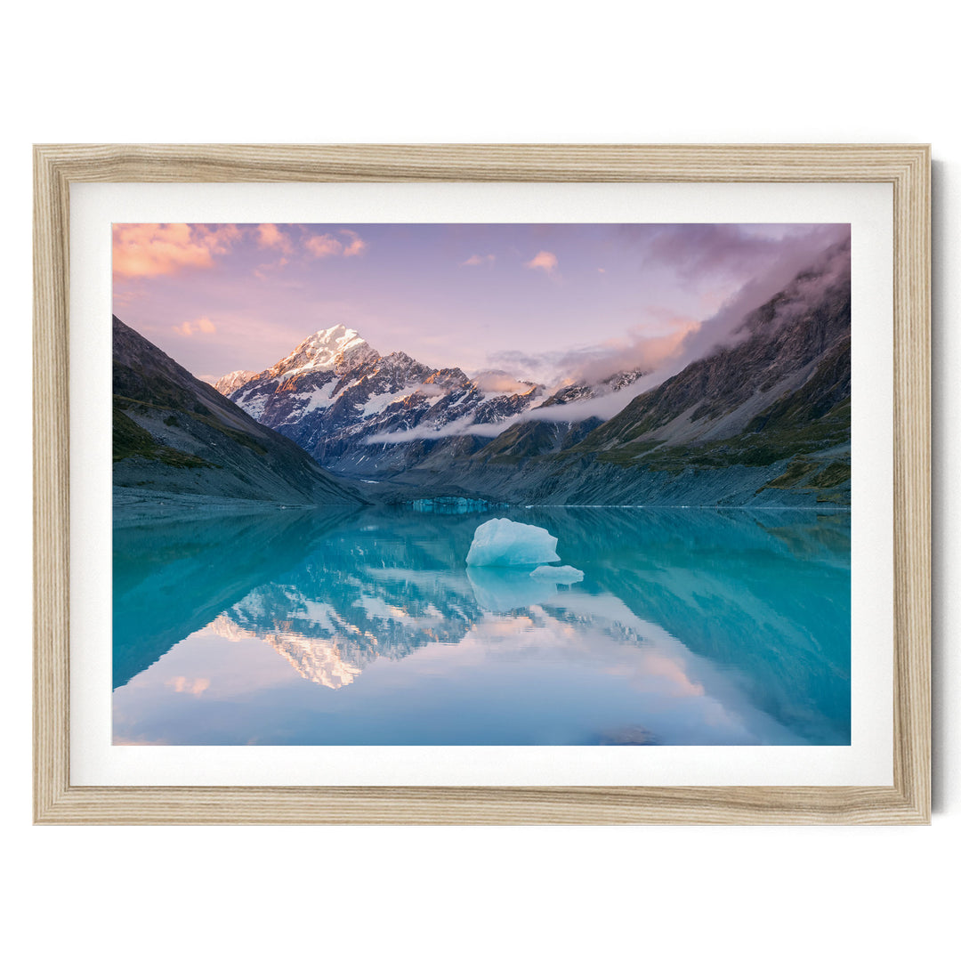 a serene mountain lake with a large ice formation in the center, surrounded by snow-capped peaks and a stunning sunset sky reflected in the calm waters.