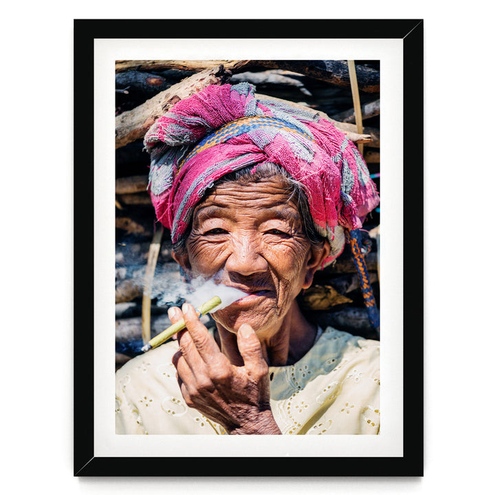 An elderly woman wearing a pink headscarf and holding a cigarette, with a pile of wood in the background.