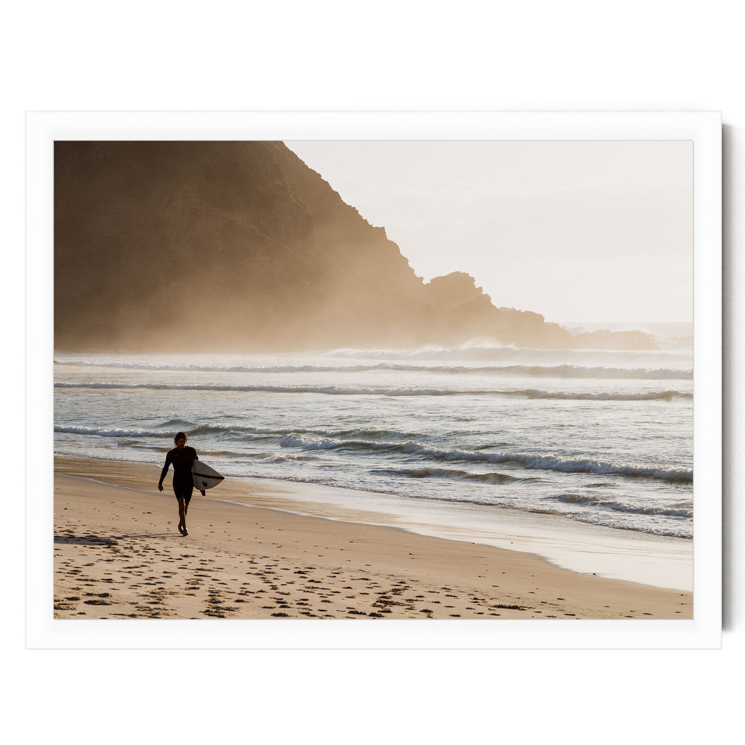 A person in a wetsuit is walking along the beach, carrying a surfboard, with a rocky cliff in the background.