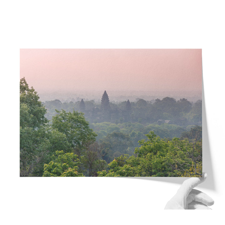 A hand is holding a framed photograph of a lush, green forest with a temple visible in the distance.