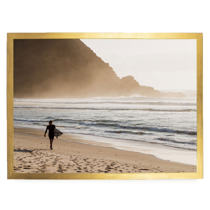 A surfer walks along the beach, carrying a surfboard, with a rocky cliff and misty mountains in the background.
