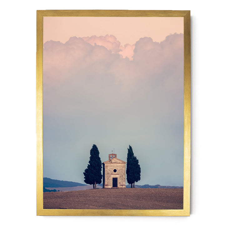A small stone church with a bell tower sits in a field, surrounded by two tall cypress trees. The sky above is filled with clouds, creating a serene and peaceful atmosphere.