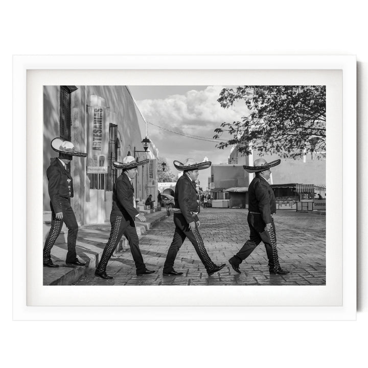 A group of men wearing sombreros walking down a street, with a building and trees in the background.