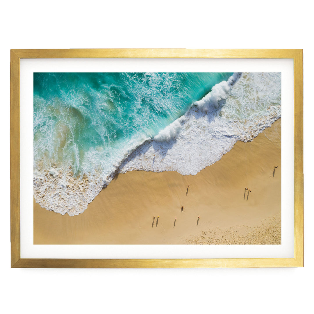 The image is a framed photograph of a beach scene, featuring a large wave crashing onto the sandy shore and people walking along the beach.