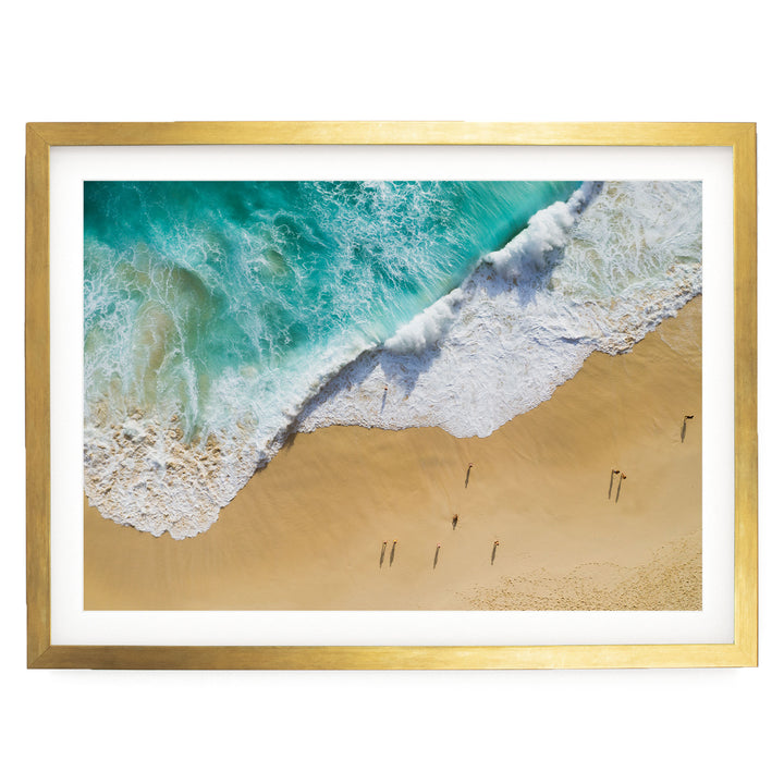 The image is a framed photograph of a beach scene, featuring a large wave crashing onto the sandy shore and people walking along the beach.