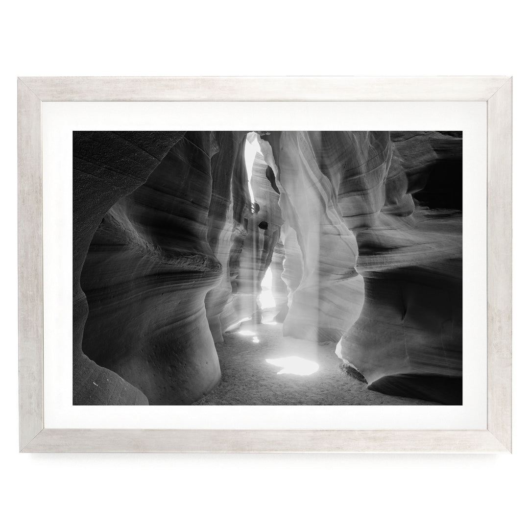 A black and white photograph of a narrow passage in a canyon, with sunlight streaming through the opening and casting shadows on the walls.