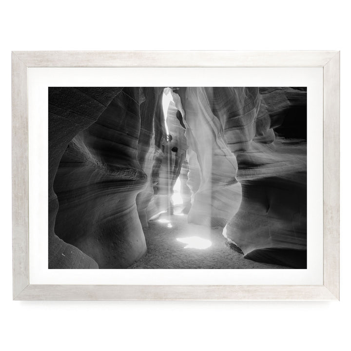 A black and white photograph of a narrow passage in a canyon, with sunlight streaming through the opening and casting shadows on the walls.