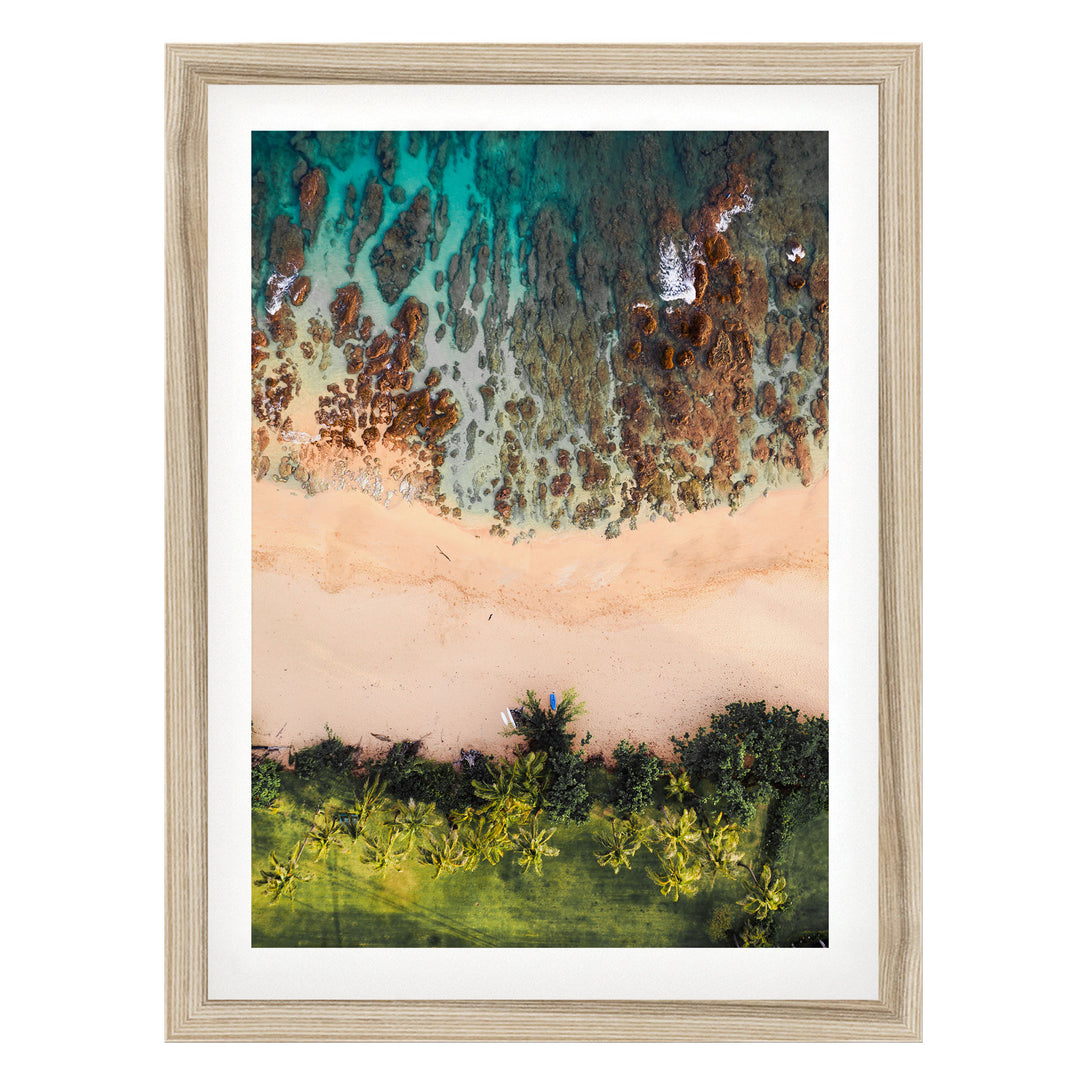 The image is a framed aerial view of a beach with a mix of sandy and rocky areas, surrounded by lush greenery and palm trees.