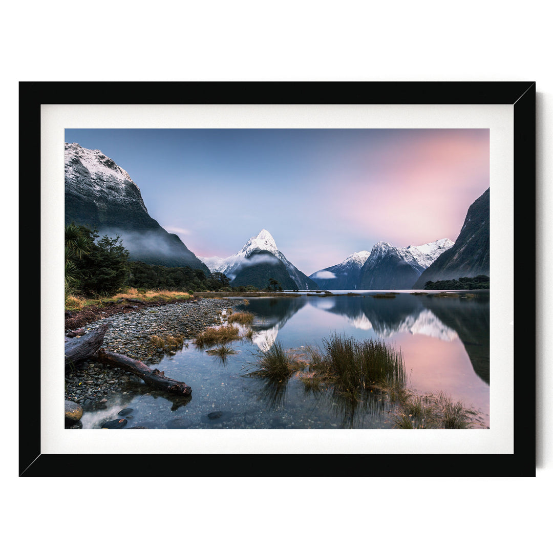 A serene landscape featuring a mountain range reflected in a calm lake, with a clear blue sky and a pinkish-orange sunset.