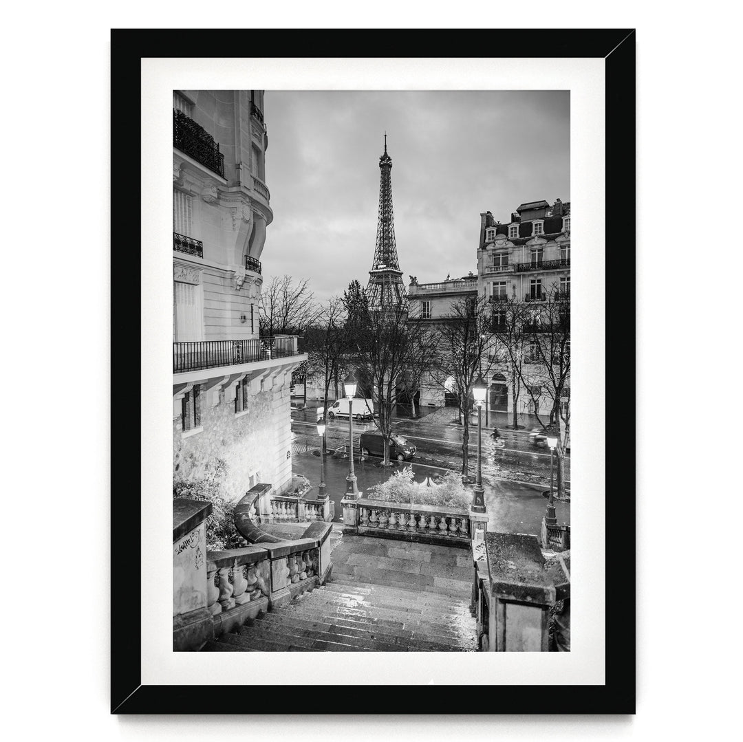 A black and white photograph of the Eiffel Tower in Paris, France, with a street scene in the foreground featuring ornate buildings and a wet, rainy atmosphere.