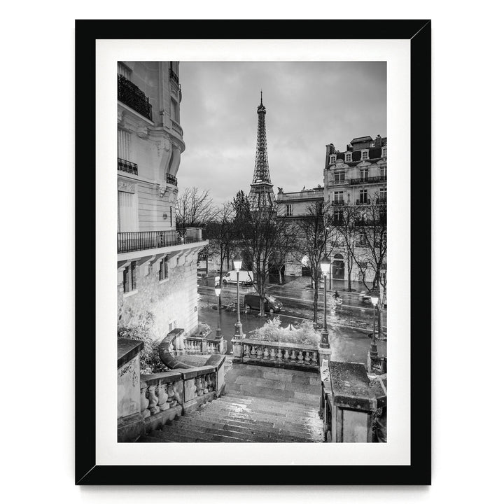 A black and white photograph of the Eiffel Tower in Paris, France, with a street scene in the foreground featuring ornate buildings and a wet, rainy atmosphere.