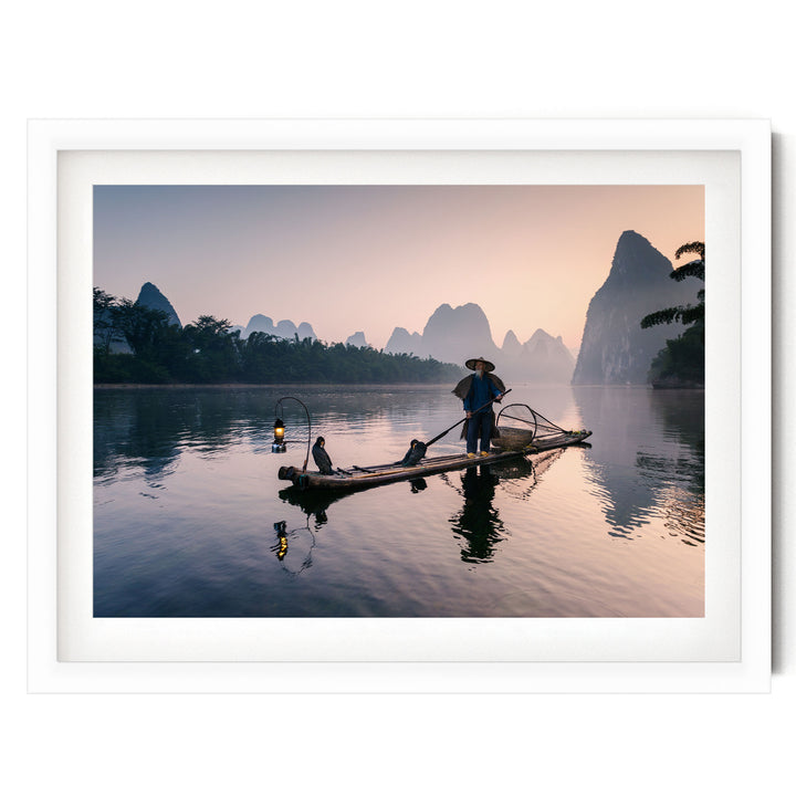 A person stands on a long wooden boat, paddling through a serene lake at sunset, with mountains in the background.