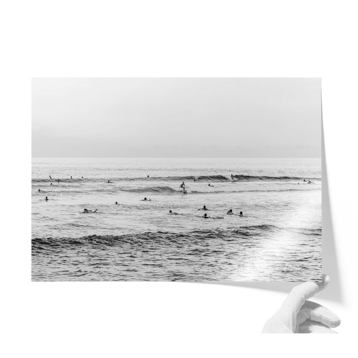 A black and white photograph of a group of people swimming and surfing in the ocean, with waves visible in the background.