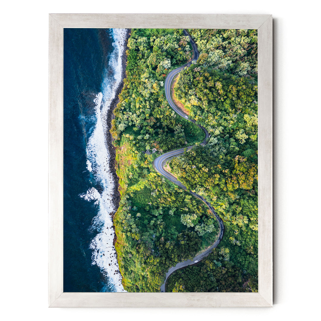The image is an aerial view of a winding road cutting through a lush, green forest. The road curves gently around the edge of a cliff, with the ocean visible on the left side. The surrounding landscape is dense with trees and vegetation, creating a serene and picturesque scene.