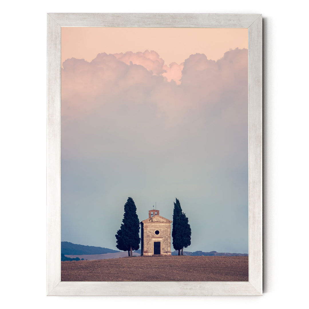 A small stone church with a bell tower sits in a field, surrounded by trees, with a cloudy sky in the background.