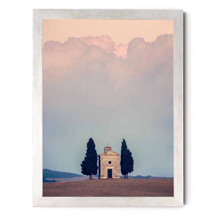 A small stone church with a bell tower sits in a field, surrounded by trees, with a cloudy sky in the background.