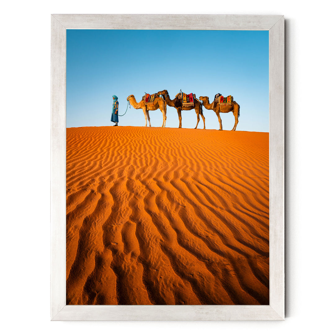 A person in a blue robe leads three camels across a vast, undulating sand dune in a desert landscape.