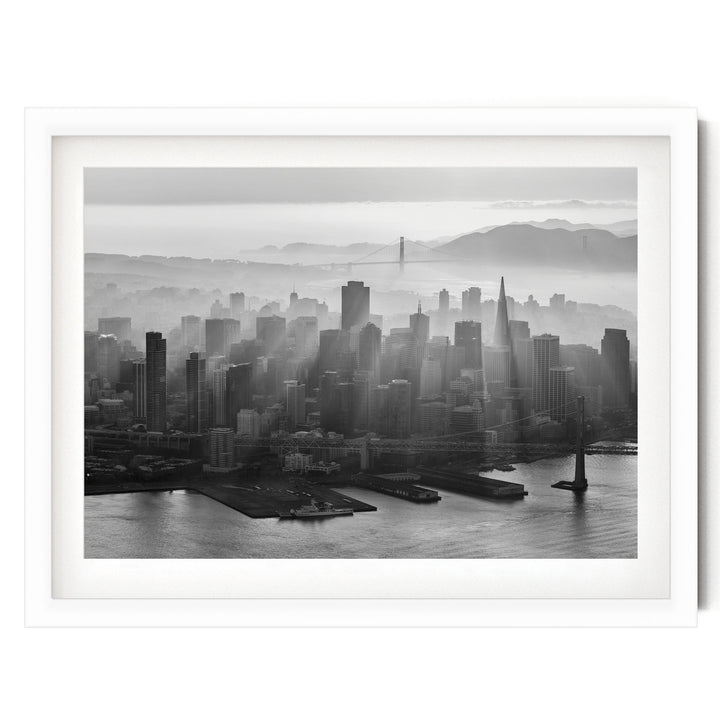 A black and white aerial view of a densely populated city skyline, with tall buildings and skyscrapers stretching out across the frame. The city is situated near a body of water, with a bridge visible in the distance. The sky above the city is hazy, creating a dramatic and atmospheric effect.