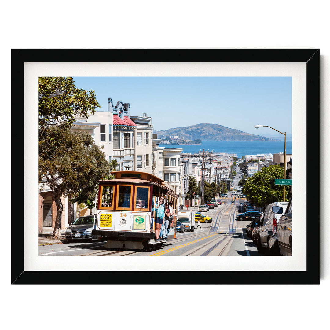 A cable car is traveling down a street lined with buildings and trees, with a mountain visible in the distance.