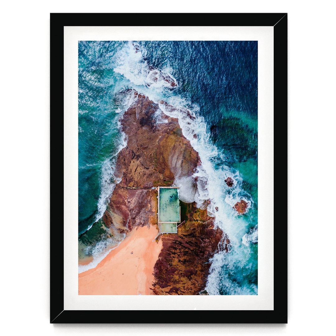The image is an aerial view of a beach with a swimming pool, surrounded by rocky cliffs and crashing waves.