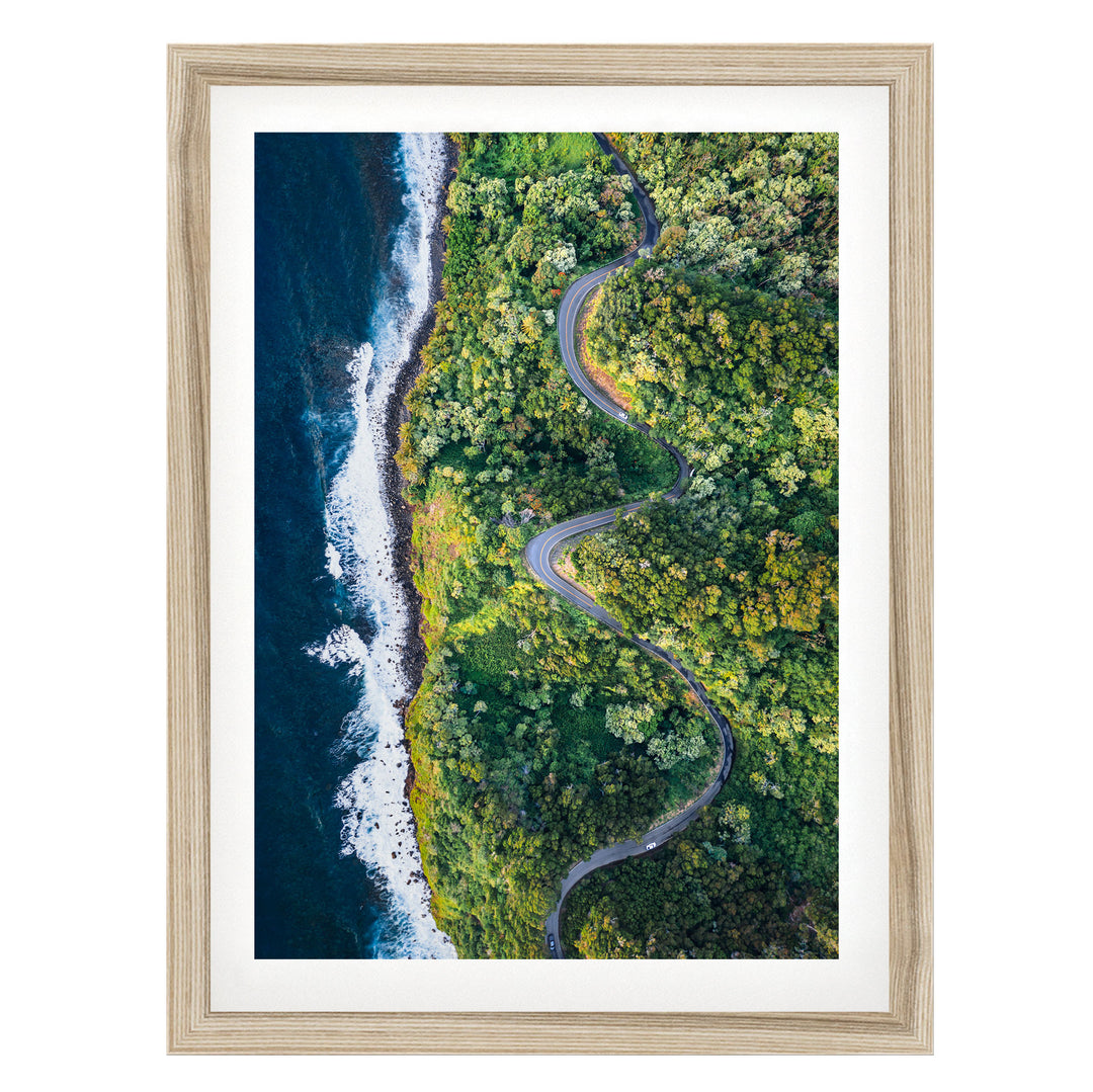 An aerial view of a winding road cutting through a lush, green forest, with the ocean visible on the left side.