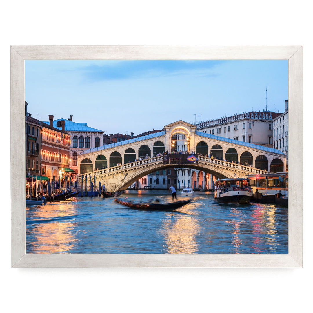 a picturesque scene of the Rialto Bridge in Venice, Italy, with its distinctive white stone arch spanning the Grand Canal. The bridge is illuminated by warm lights, creating a romantic atmosphere. The canal is filled with boats, adding to the lively ambiance of the scene. The image is framed, suggesting it is a part of a larger collection or a printed photograph.