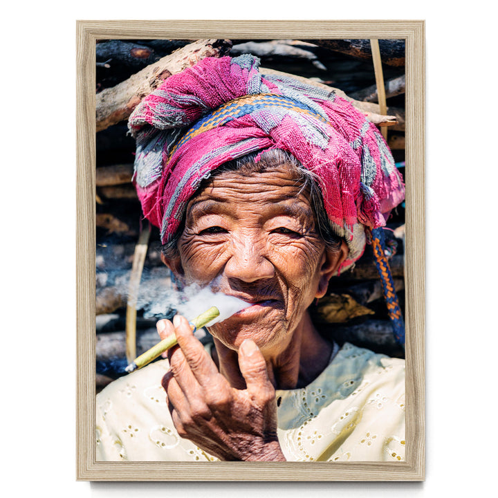 An elderly woman wearing a colorful headscarf and holding a lit cigarette, with a pile of wood in the background.