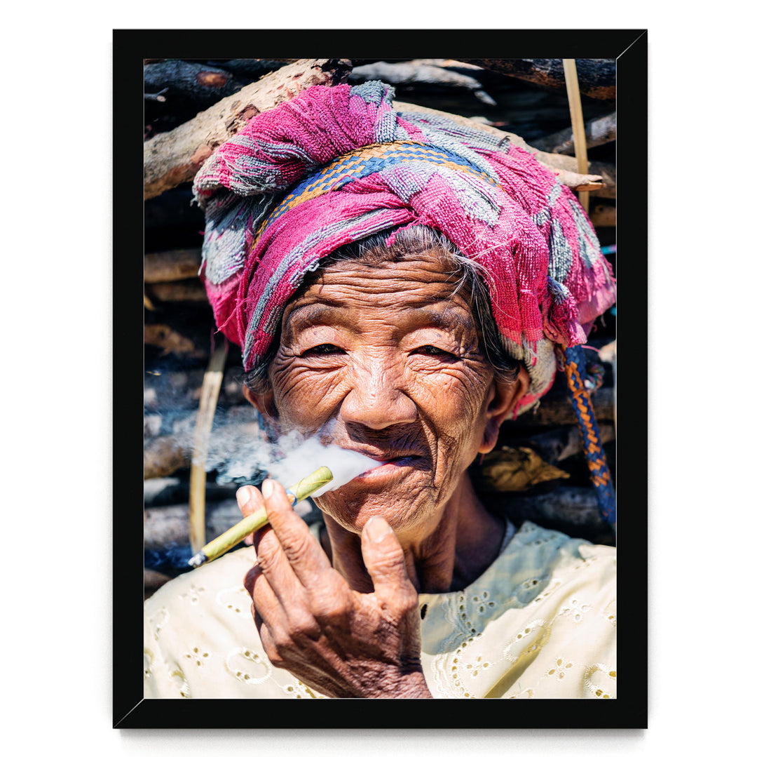 An elderly woman wearing a pink headscarf and a white blouse is shown smoking a cigarette, with a pile of wood visible in the background.