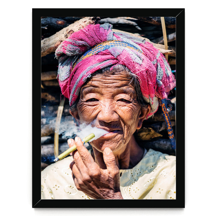 An elderly woman wearing a pink headscarf and a white blouse is shown smoking a cigarette, with a pile of wood visible in the background.
