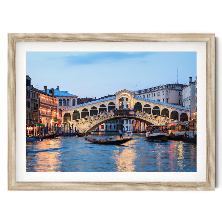 a picturesque scene of the Rialto Bridge in Venice, Italy, with its distinctive white stone arch spanning the Grand Canal. The bridge is illuminated at dusk, creating a warm and inviting atmosphere. The canal is filled with boats, adding to the lively and bustling ambiance of the scene. The image is framed and appears to be a piece of art or a photograph, showcasing the beauty and charm of this iconic landmark.