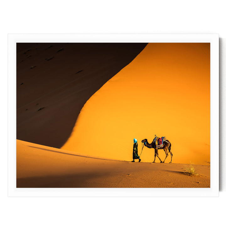 A person in traditional desert attire walks alongside a camel in a desert landscape with a large sand dune in the background.