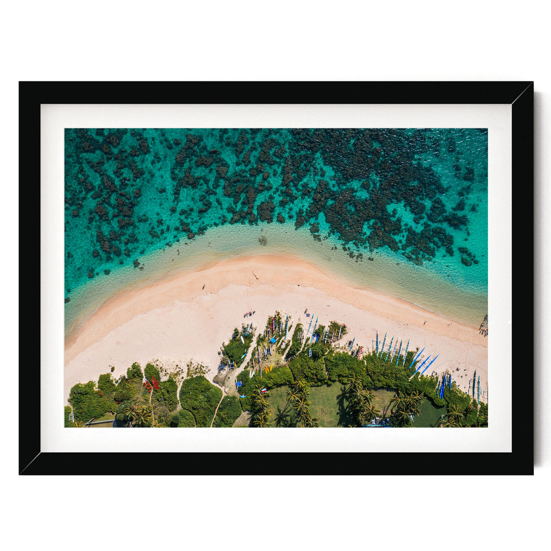 The image is an aerial view of a tropical beach with crystal clear turquoise waters, sandy shores, lush vegetation, and a few boats visible in the distance.