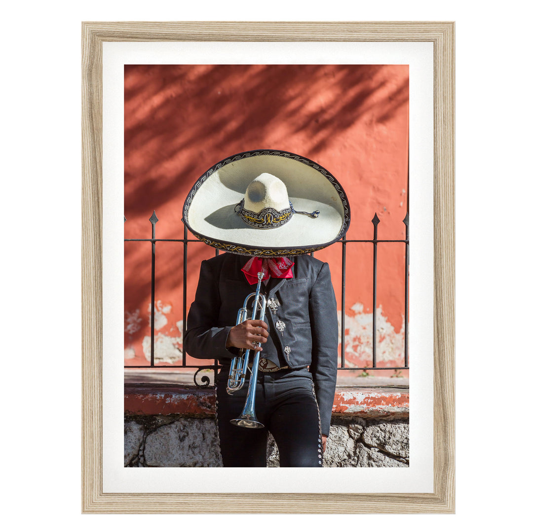 A person wearing a black jacket and a large white sombrero is playing a trumpet in front of a red wall.