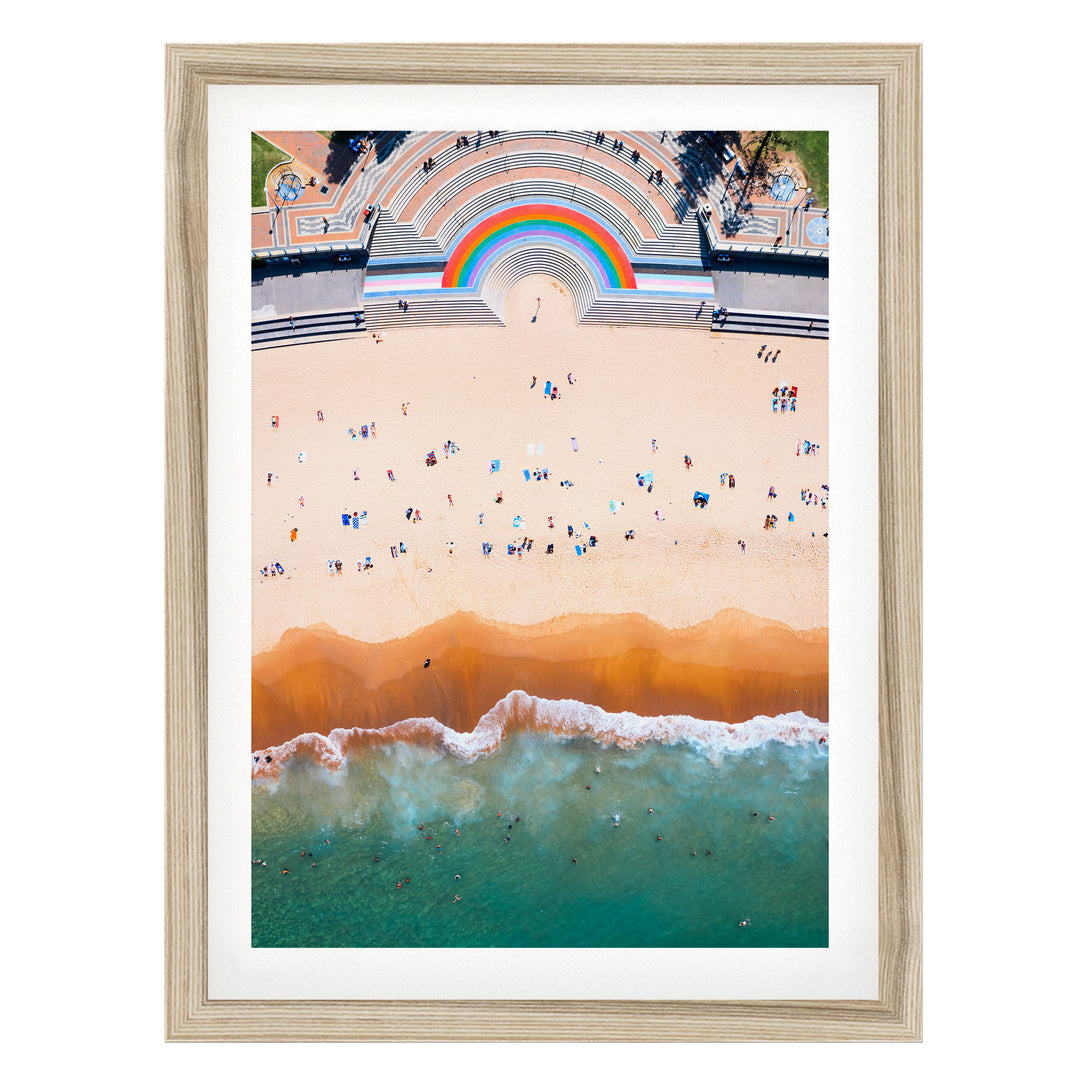 The image is an aerial view of a beach with a large crowd of people enjoying the sunny day. The beach is lined with a large, colorful arch structure, and the ocean is visible in the background.
