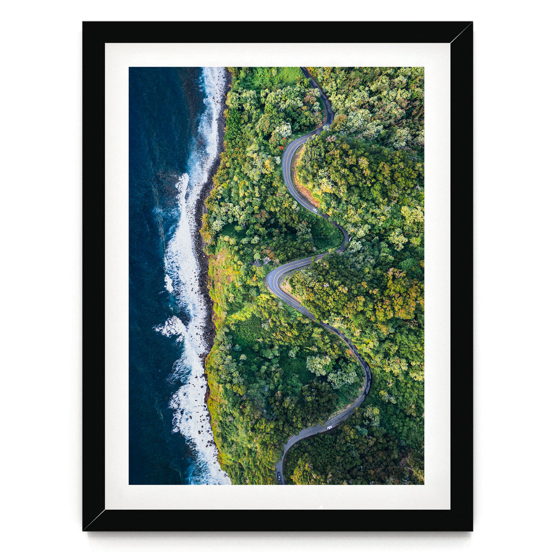 An aerial view of a winding road cutting through a lush, green forest, with the ocean visible on the left side.