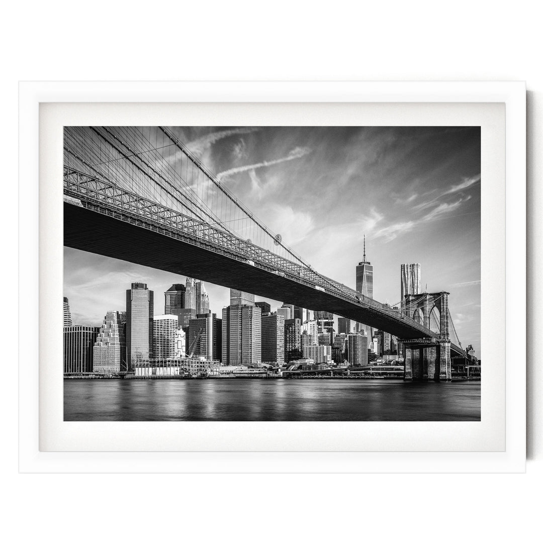 A black and white photograph of the Brooklyn Bridge spanning the East River, with the New York City skyline visible in the background.