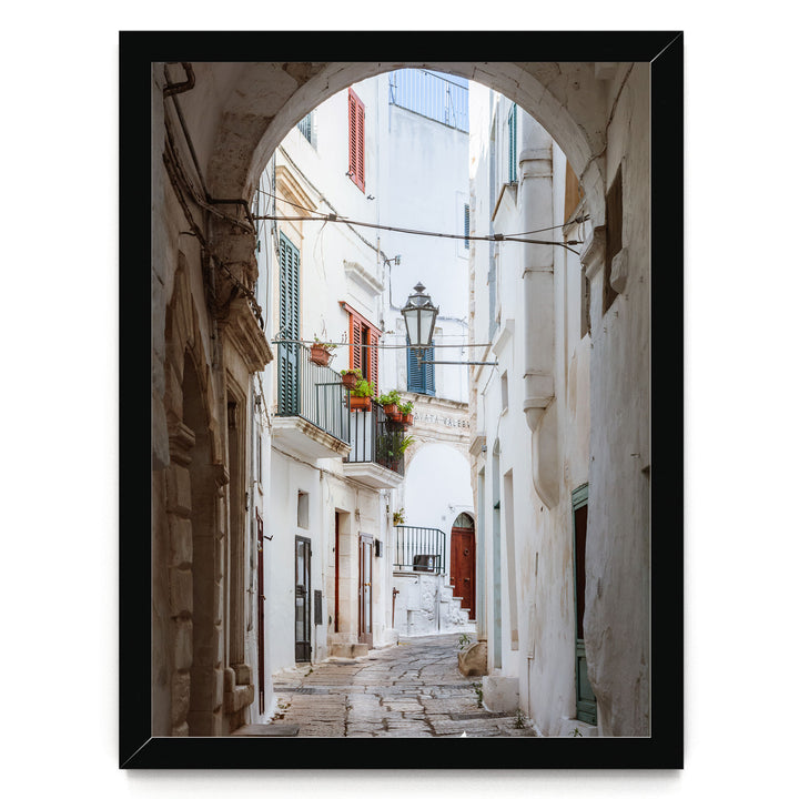 A narrow, cobblestone street lined with white buildings and colorful shutters, leading to a balcony with potted plants.